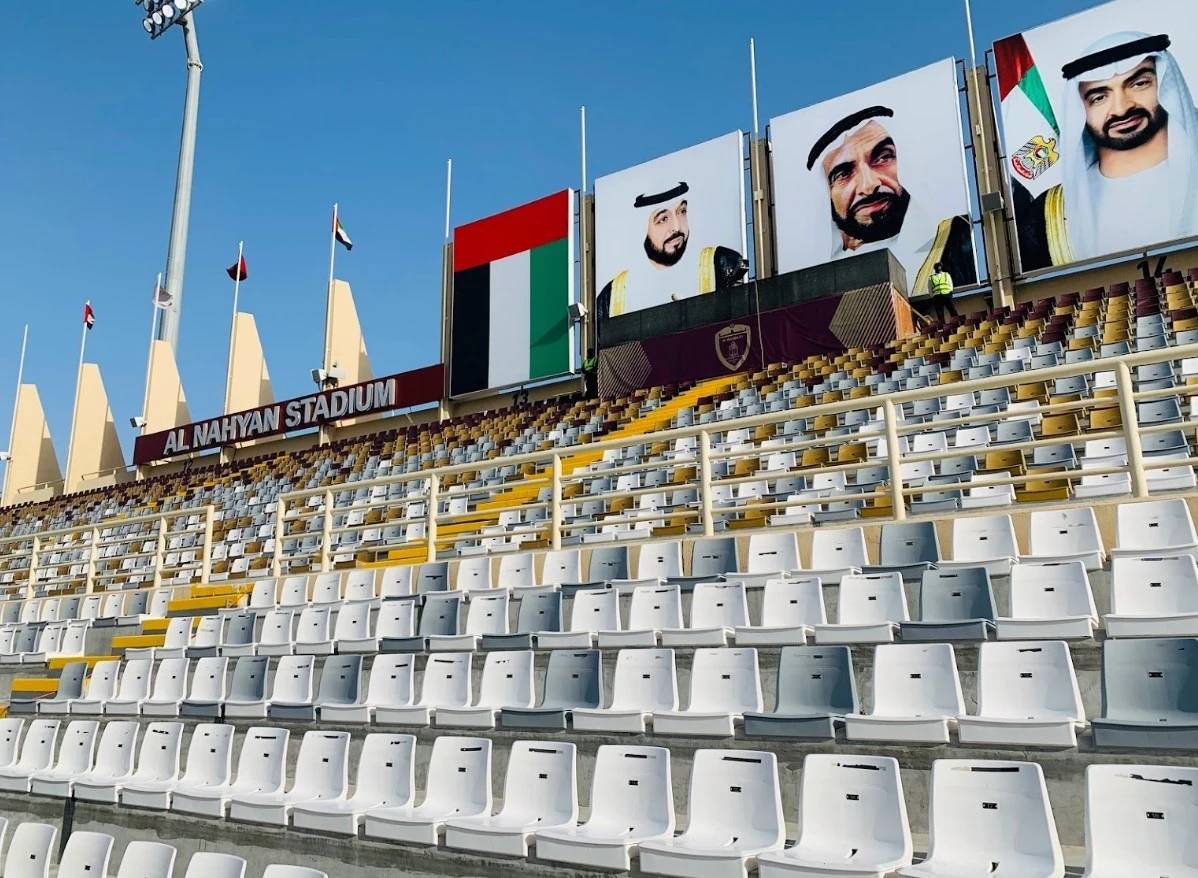 Empty seats at Al Nahyan Stadium, Abu Dhabi, with large portraits displayed.